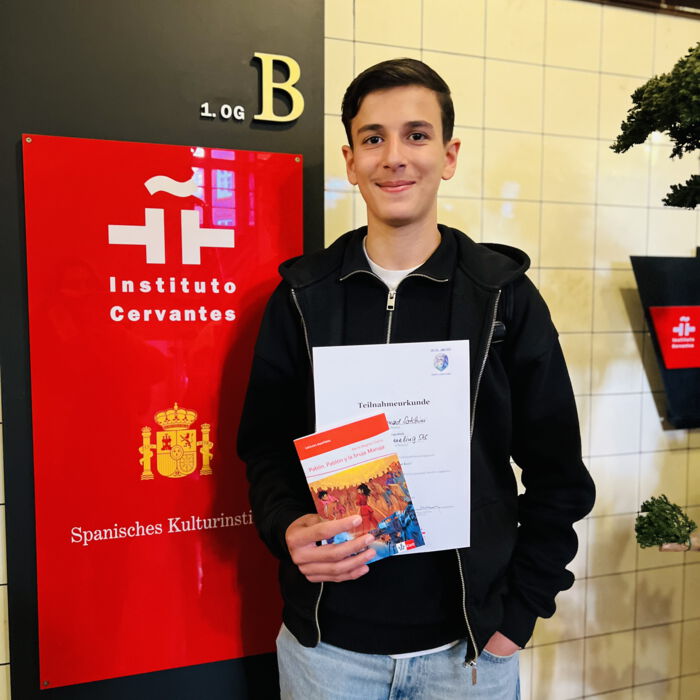 Schüler mit Urkunde und Buch vor einer Aufstellwand des Instituto Cervantes
