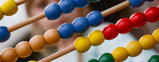 Ein Abakus mit bunten Perlen in Gelb, Blau, Grün und Rot ist im Vordergrund zu sehen. Im Hintergrund, unscharf, deutet sich eine Person an