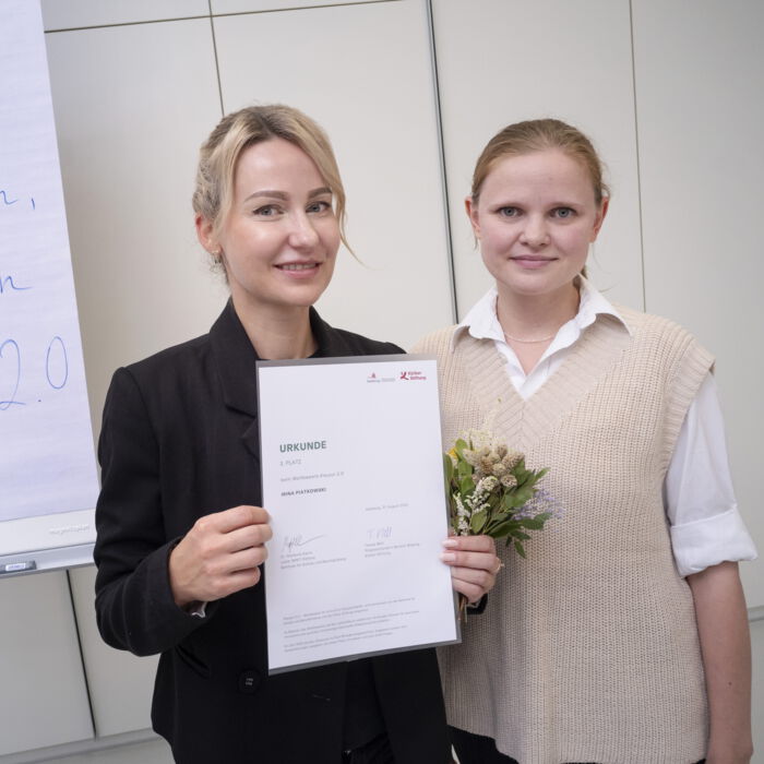 Zwei Frauen lächeln in die Kamera, eine hält eine Urkunde, die andere einen kleinen Blumenstrauß. Im Hintergrund steht ein Flipchart mit handgeschriebenem Text