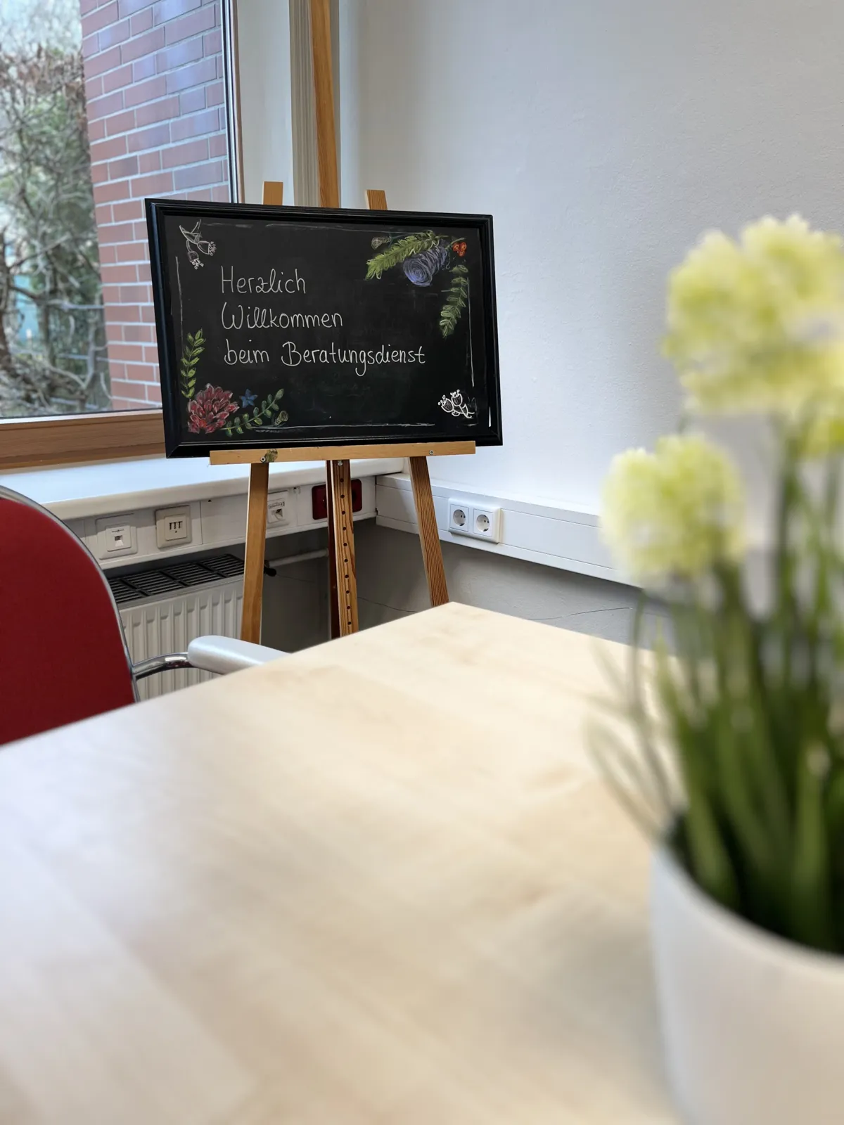 Heller Beratungsraum mit einer kleinen Tafel auf einer Staffelei, Tisch und Blumen am Fenster