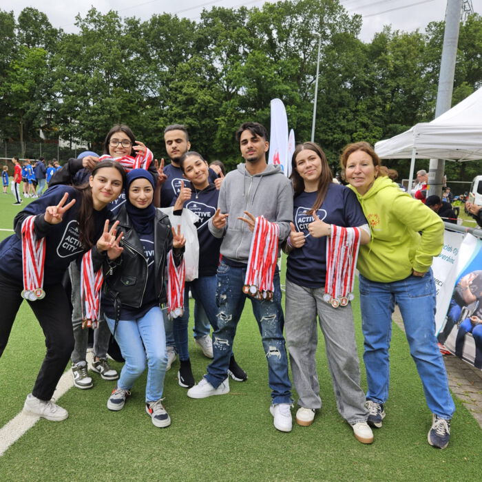Eine Gruppe junger Menschen posiert fröhlich auf einem Sportplatz mit Medaillen. Sie tragen T-Shirts mit der Aufschrift 'Active City' und zeigen Peace-Zeichen. Im Hintergrund sind ein Fußballspiel und Zuschauer zu sehen