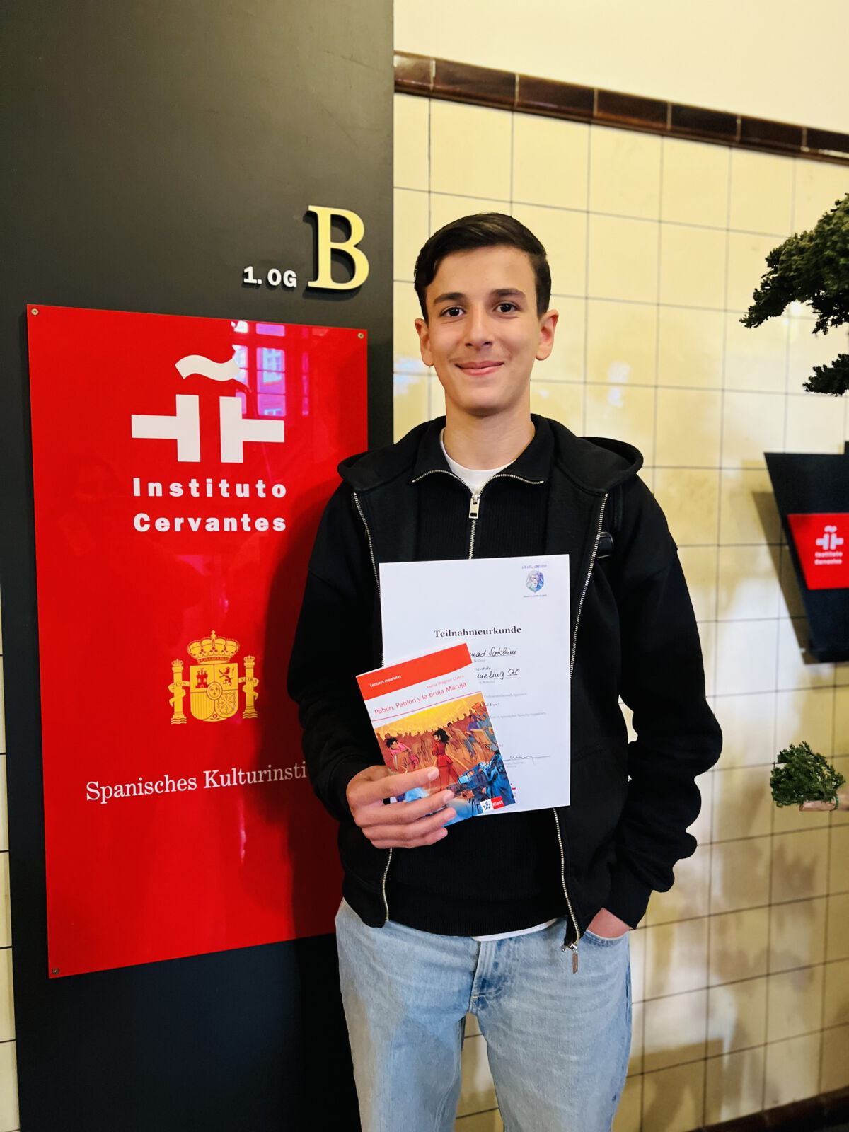 Schüler mit Urkunde und Buch vor einer Aufstellwand des Instituto Cervantes