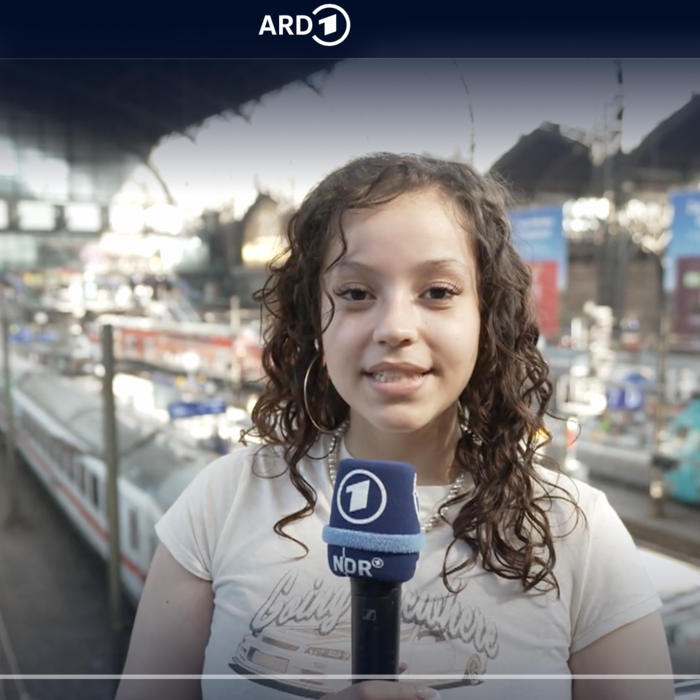 Ein junges Mädchen hält ein Mikrofon mit dem NDR-Logo und spricht in die Kamera. Im Hintergrund ist ein belebter Bahnhof mit Zügen und Gleisen zu sehen.