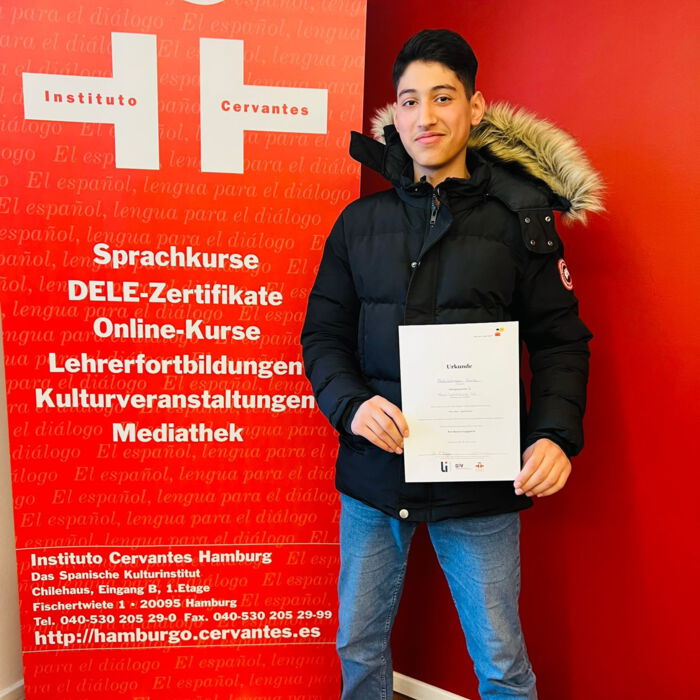 Ein Jugendlicher steht vor einem roten Banner des Instituto Cervantes Hamburg und hält stolz ein Zertifikat in der Hand