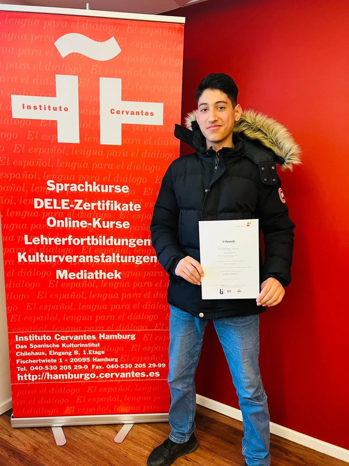 Ein Jugendlicher steht vor einem roten Banner des Instituto Cervantes Hamburg und hält stolz ein Zertifikat in der Hand