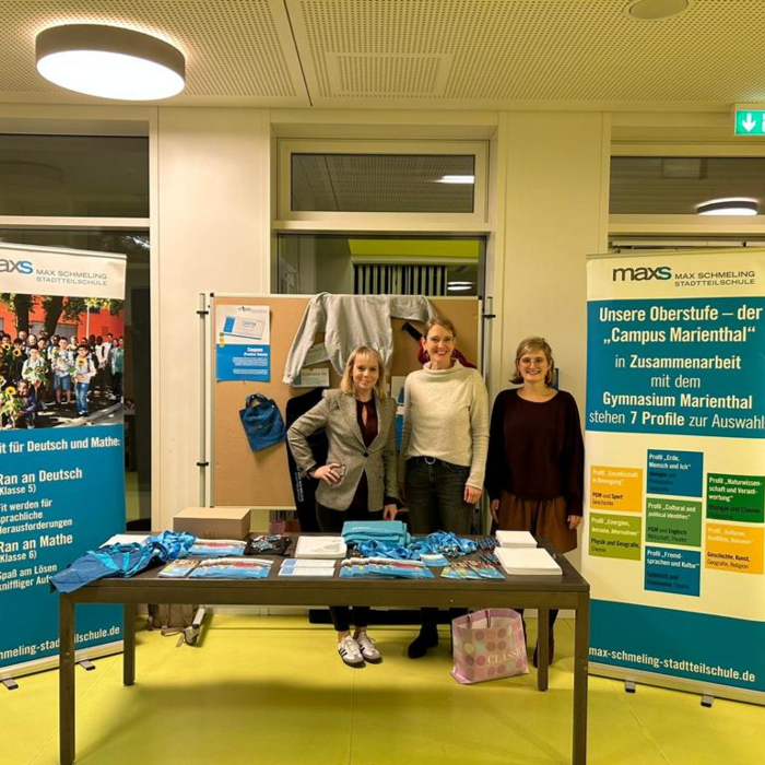 Drei Frauen stehen an einem Informationsstand der Max-Schmeling-Stadtteilschule, flankiert von Roll-Ups mit Informationen zu Schulprogrammen. Auf dem Tisch liegen Flyer und kleine Geschenke aus