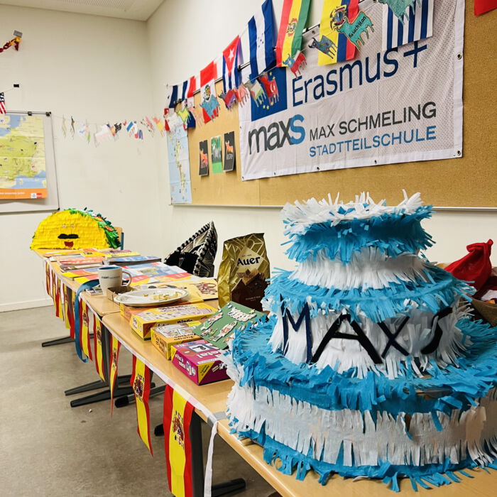 Ein Raum mit Erasmus+-Banner der Max-Schmeling-Stadtteilschule. Auf einem Tisch stehen Spiele, Snacks und eine blau-weiße Piñata mit 'MAX'. Flaggen und Zeichnungen dekorieren den Raum
