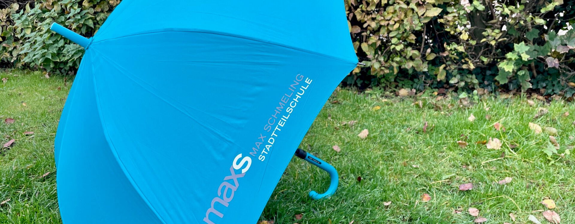 Ein blauer Regenschirm mit dem Logo der Max-Schmeling-Stadtteilschule liegt geöffnet auf einer grünen Wiese vor einer Hecke.