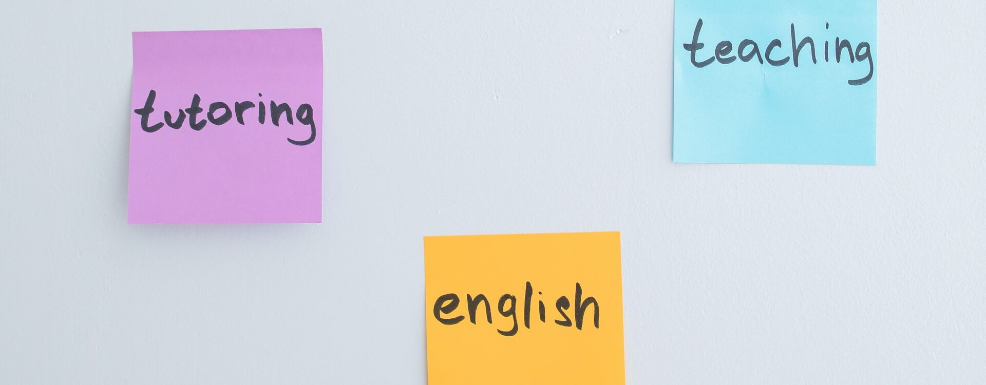 Drei Post-its an einer Wand, auf denen 'tutoring', 'english' und 'teaching' geschrieben steht, jeweils auf farbigen Hintergründen in Lila, Gelb und Blau