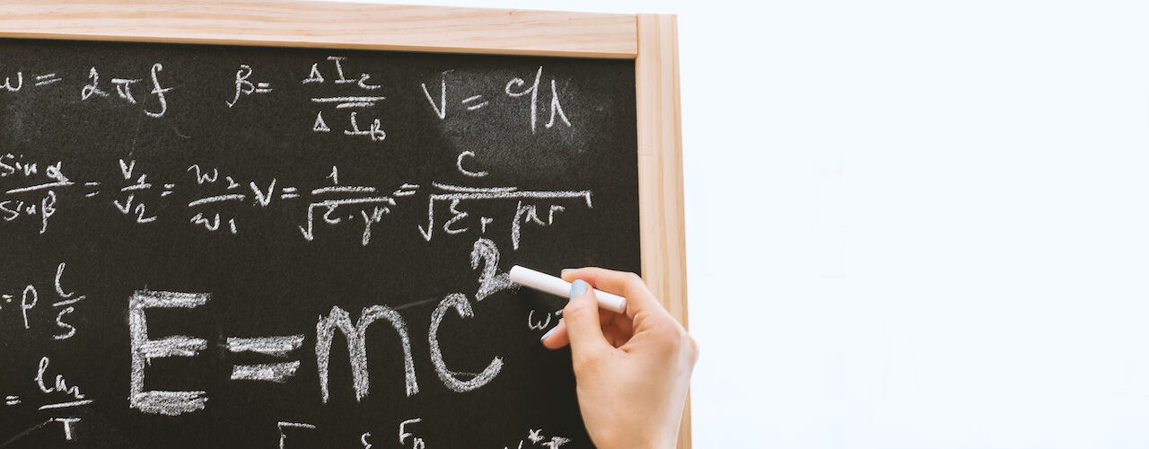 Eine Hand schreibt mit Kreide die Formel „E=mc²“ auf eine Tafel, die mit weiteren physikalischen Formeln gefüllt ist.