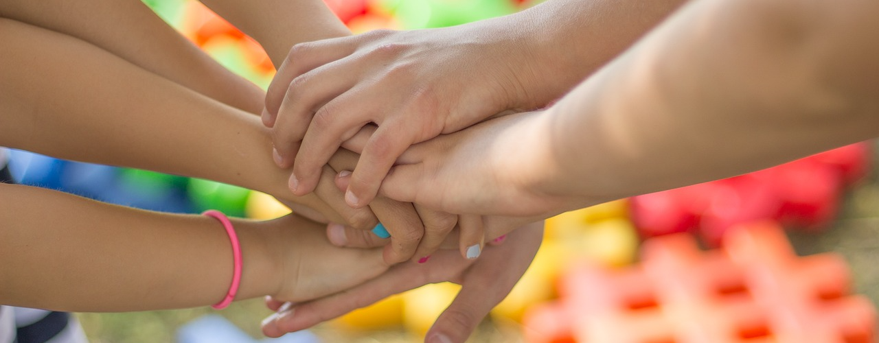 Mehrere Hände von Kindern und Erwachsenen, die übereinander gestapelt sind, vor einem bunten Hintergrund aus Spielbausteinen