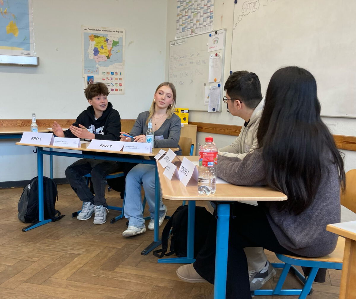 Vier Schüler*innen sitzen an Tischen in einem Klassenzimmer und diskutieren engagiert. Vor ihnen stehen Namensschilder