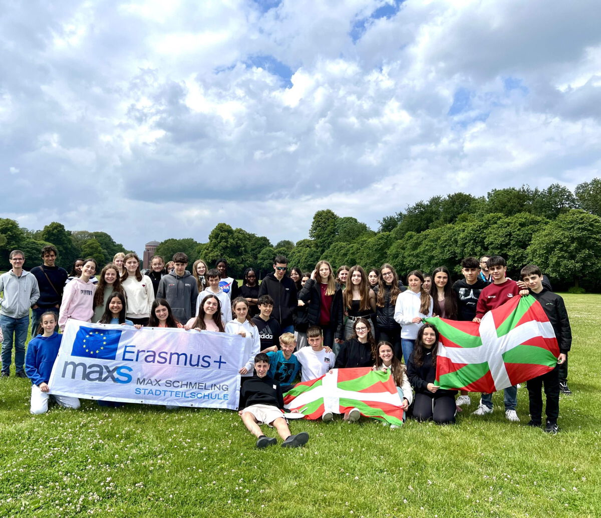 Eine große Gruppe von Schüler*innen steht auf einer Wiese unter einem bewölkten Himmel. Sie halten ein Erasmus+-Banner der Max-Schmeling-Stadtteilschule sowie baskische Flaggen