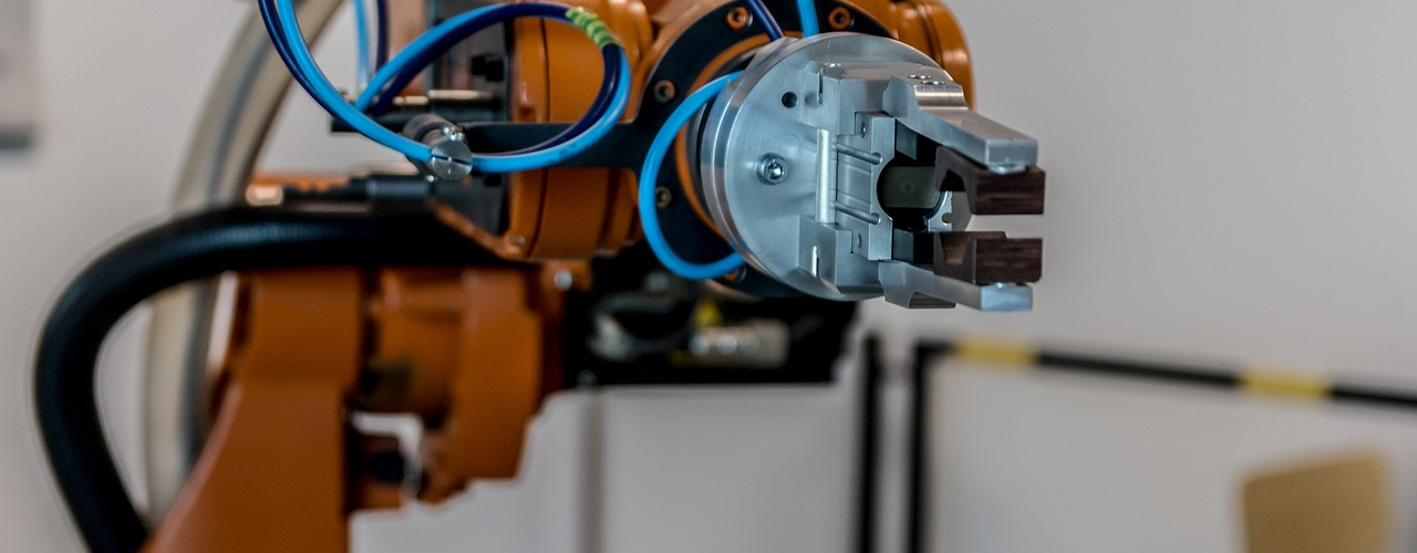 Ein Industrieroboterarm mit blauer Verkabelung und einer Greifvorrichtung im Fokus. Der Hintergrund ist unscharf, was den Roboter in den Mittelpunkt rückt.